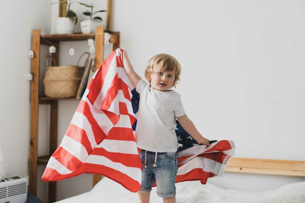 Best places to hang a custom flag in your home - flags usa - little boy holds american flag as he jumps on the bed